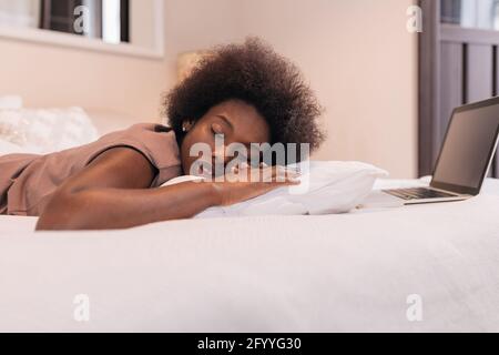 Seitenansicht der müden jungen afroamerikanischen Freiberuflerin mit Lockige dunkle Luft in legerer Kleidung, die auf dem Bett und liegt Schlafen nach der Distanz Arbeit auf dem Schoß Stockfoto