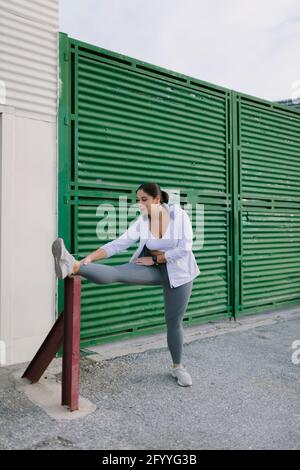 Ganzkörper aus flexibler junger, ethnischer Sportlerin in aktiven Stretching-Beinen nach dem Joggen auf der Straße der Stadt Stockfoto