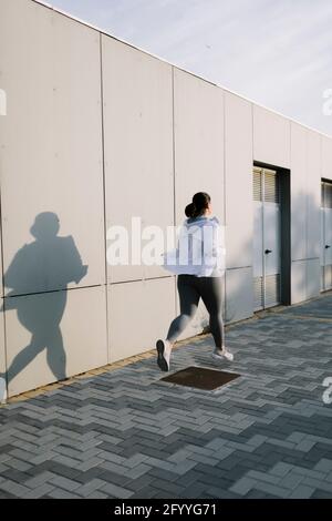 Rückansicht einer nicht erkennbaren, entschlossenen jungen und großen Sportlerin Mit dunklem Haar in Sportkleidung, die auf der Stadtstraße in der Nähe läuft Betonwand Stockfoto