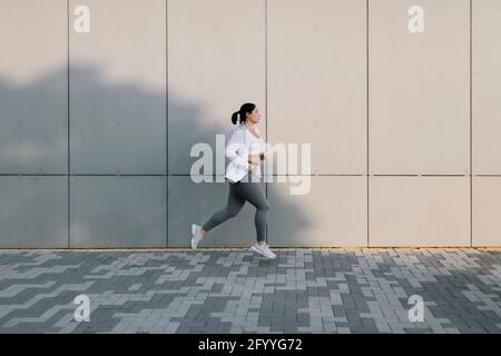Seitenansicht der entschlossenen jungen und großen Sportlerin mit Dunkles Haar in Sportkleidung, das auf der Stadtstraße in der Nähe von Beton läuft Wand Stockfoto