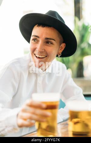 Fröhlicher, stilvoller Tomboy mit Tattoos, Hut und weißem Hemd Bier aus Glas im Café trinken Stockfoto