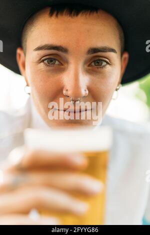 Positiver Tomboy mit Tattoos mit Hut und weißem Hemd Bier aus Glas im Café Stockfoto