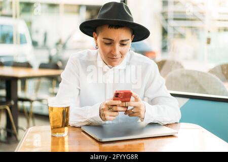 Konzentrierter Tomboy in Hut sitzend am Tisch mit Laptop und Ein Glas Bier beim Scrollen auf dem Mobiltelefon Stockfoto
