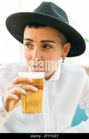 Positiver Tomboy mit Tattoos mit Hut und weißem Hemd Bier aus Glas im Café Stockfoto