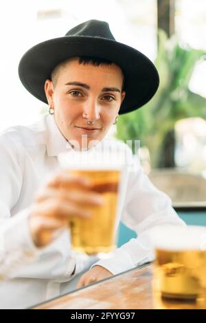 Positiver Tomboy mit Tattoos mit Hut und weißem Hemd Bier aus Glas im Café Stockfoto