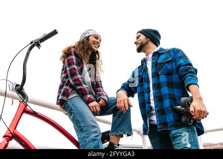 Von unten junge glückliche Freunde lächeln und sprechen in der Nähe Metallgeländer und Fahrrad tagsüber Stockfoto