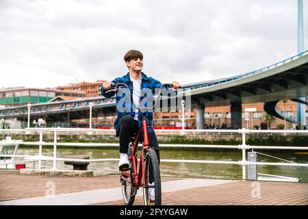 Ganzkörper von jungen Mann in stilvollen Sneakers Fahrrad fahren Und unter dem wolkigen Himmel in der Stadt wegschauen Stockfoto