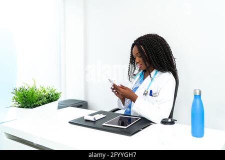 Positive afroamerikanische Ärztin beim Surfen auf dem Handy während Arbeiten im medizinischen Krankenhaus am Tisch mit Tablet Stockfoto