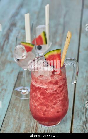 Glaskrug mit kaltem Wassermelone Gin Cocktail in der Nähe von Kelchen platziert Mit Eis und Früchten auf Holztisch Stockfoto