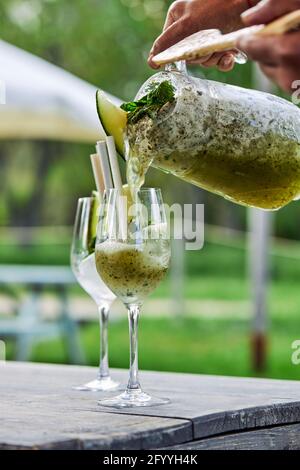 Unkenntliche Person, die frischen Rum-Cocktail mit Melone und Minze gießt Im Glas Becher auf dem Tisch am Sommertag im Hof Stockfoto