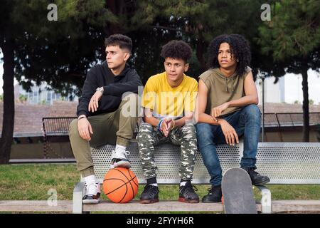 Ganzer Körper von multirassischen männlichen Freunden sitzen auf Bank mit Basketball und Skateboard und aufmerksam wegschauen Stockfoto