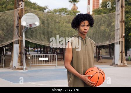 Afroamerikanischer Teenager-Spieler mit Ball, der gegen das Netz steht Sportplatz und Blick auf die Kamera Stockfoto