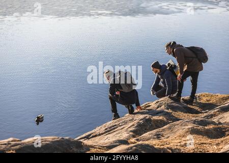 Seitenansicht von jungen männlichen Reisenden in warmer Oberbekleidung und Rucksäcke sitzen auf Geisterhacken auf felsigen Klippen und bewundern niedlich mallard schwimmt auf dem See Stockfoto