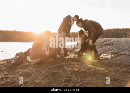 Voller Körper von fröhlichen jungen, vielfältigen Reisenden Freunden in warmen Kleidung, die am Seeufer sitzt und während des Campens Feuer anzündet Im Herbst Wald auf sonnig Stockfoto