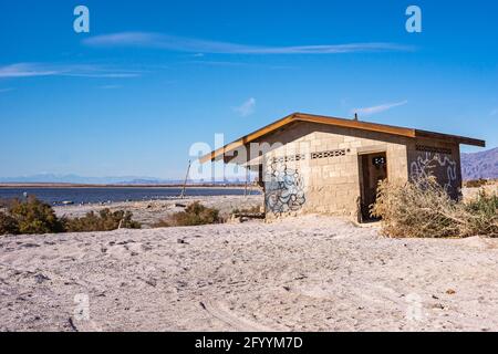 USA, CA, Salton Sea - 28. Dezember 2012: Ein echtes Gebäude aus braunem Stein für die Toilette am Strand von Niland Marina unter blauem Himmel. Weißer grauer Sand und das Meer an Stockfoto