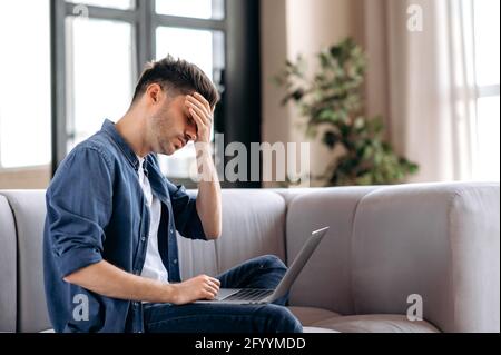 Müde stressige junge kaukasische bärtige Mann sitzt auf dem Sofa im Wohnzimmer, in legerer Denim-Kleidung, mit Laptop, Freiberufler überarbeitet mit entfernter Arbeit, Kopfschmerzen, braucht Ruhe Stockfoto