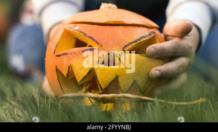 Bodenwinkelaufnahme. Nahaufnahme der Hände eines Kindes, die geschnitzten Kürbis, Jack-o-Laterne, in das Gras, vor die Kamera setzen Stockfoto