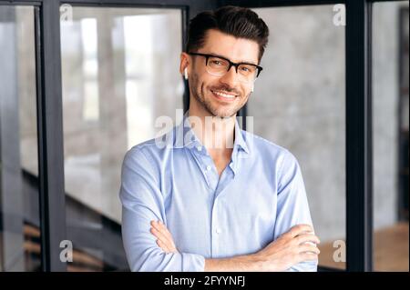 Porträt eines fröhlichen, attraktiven, selbstbewussten jungen Geschäftsmanns oder Geschäftsführers, der Brille und Kopfhörer trägt, ein formelles Hemd trägt, im Büro steht, die Kamera anschaut, die Arme gekreuzt, Freundlich lächelnd Stockfoto