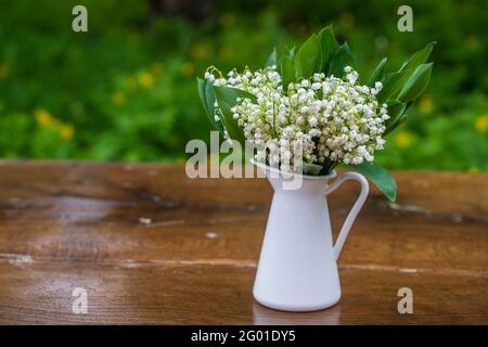 Schönes Bouquet von Lilien des Tals in einer weißen emaillierten Vase auf dem Holztisch im Garten, aus nächster Nähe Stockfoto
