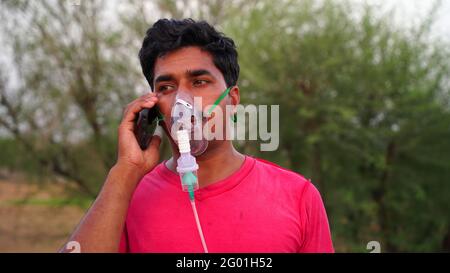 Junger Mann, der mit der Covid-19-Krankheit infiziert ist. Patient inhaliert Sauerstoff trägt Maske und Anrufe auf Handy. Flüssiger Sauerstoff-Flow in Rajasthan, Indien. Stockfoto
