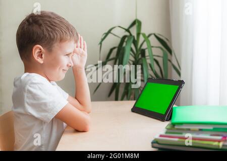 Lächelnder Schuljunge sitzt am Schreibtisch und schaut auf das Tablet mit grünem Bildschirm. Ein Kind zieht an der Hand, um zu antworten. Online-Unterricht aus der Ferne. Grüne Chroma-Taste auf dem Monitor. Seitenansicht Stockfoto