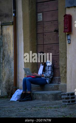 Arezzo, Italien, 28. Mai 2021, junge Studentin mit langen Haaren im Gesicht, die ihren Laptop auf der Steinstufe sitzend benutzt Stockfoto