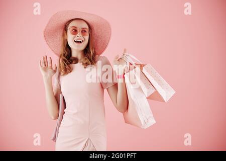Porträt einer aufgeregten jungen Frau mit Hut und Sonnenbrille in der Hand Einkaufstaschen und winken mit der Hand Stockfoto