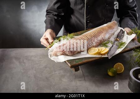 Crop gesichtslosen Koch hält Schneidebrett mit frischem rohem Fisch Filet, Kräuter und Zitrone, während Sie köstliche Meeresfrüchte zubereiten In der Küche Stockfoto