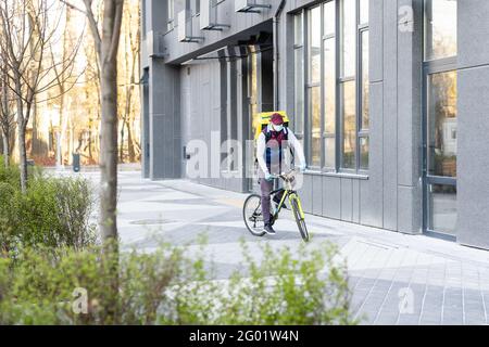 Junger Kurier in medizinischer Maske liefert Lebensmittel mit gelbem Thermalrucksack, fährt mit dem Fahrrad in der Stadt. Konzept für den Lieferservice von Lebensmitteln Stockfoto