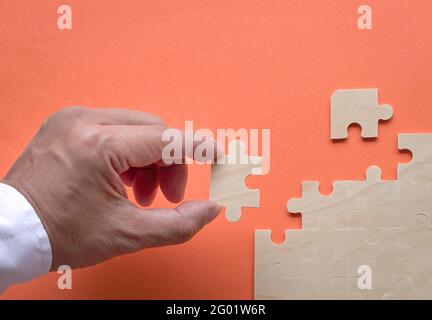 Die Hand des Mannes fügt ein Puzzleteil in das Jigzaw-Puzzle auf der orangefarbenen Tischplatte ein. Speicherplatz kopieren. Stockfoto