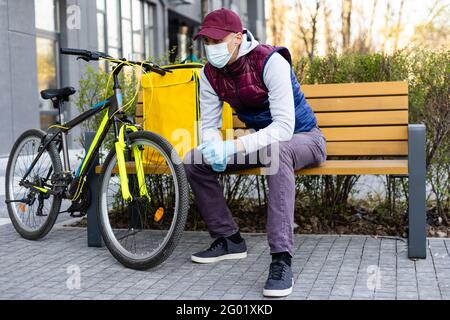 Junger Kurier in medizinischer Maske liefert Lebensmittel mit gelbem Thermalrucksack, fährt mit dem Fahrrad in der Stadt. Konzept für den Lieferservice von Lebensmitteln Stockfoto