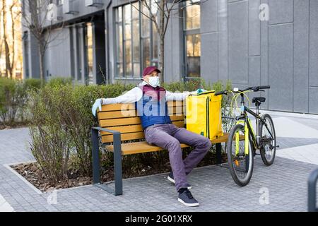 Junger Kurier in medizinischer Maske liefert Lebensmittel mit gelbem Thermalrucksack, fährt mit dem Fahrrad in der Stadt. Konzept für den Lieferservice von Lebensmitteln Stockfoto