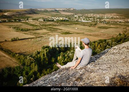 Rückansicht eines jungen Mannes, der Tee oder Kaffee in den Bergen trinkt. Reisender Mann mit Bart hält in den Händen eine Tasse Tee, nach dem Wandern. Reisen, Lifestyle, VA Stockfoto
