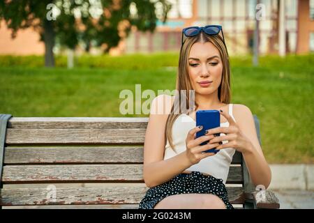 Die Frau in der Stadt sitzt im Sommer auf der Bank, hält Smartphone, liest und schreibt im Chat eine Nachricht, trägt Sonnenbrillen und Sommerkleidung. Hochwertige Fotos Stockfoto