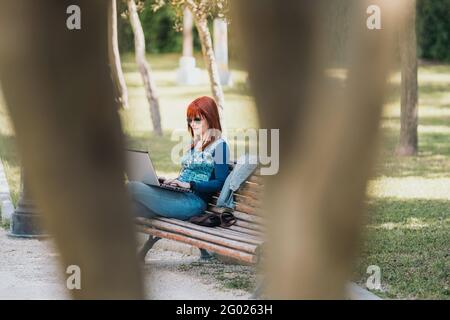 Eine rothaarige Frau, die an einem sonnigen Tag auf einer Parkbank mit einem Laptop sitzt und Kopfhörer durch die Bäume sieht. Weitschuss. Stockfoto