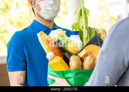 Asiatischer Lieferer in blauer Uniform mit Gesichtsmaske, während er Lebensmittel an den Kunden zu Hause liefert, Lebensmittelzustellung in der Zeit des Pandemiekonzepts Stockfoto