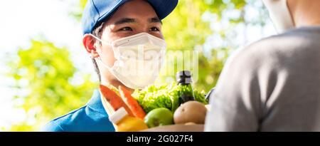 Hygienischer asiatischer Lieferer, der Gesichtsmaske trägt, während er Lebensmittel an den Kunden zu Hause liefert, und die Lieferung von Lebensmitteln in der Zeit des Pandemiekonzepts Stockfoto