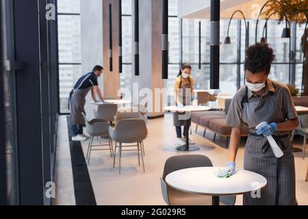 Weitwinkelaufnahme von maskierten Arbeitern beim Reinigen von Tischen im Café-Innenraum, covid Sicherheitskonzept, Kopierraum Stockfoto
