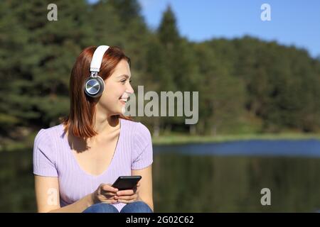 Fröhliche Frau, die mit dem Telefon und kabellosen Kopfhörern Musik hört Blick auf Side in einem See Stockfoto