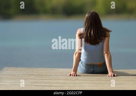 Rückansicht Porträt einer Frau, die auf einem Pier sitzt Betrachten Sie den See allein in den Sommerferien Stockfoto