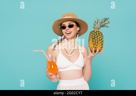 Fröhliche Frau in Sonnenbrille, die Ananas und Cocktail isoliert hält Blau Stockfoto