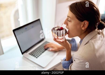 Eine junge Geschäftsfrau, die einen Laptop benutzt, während sie in einer angenehmen Atmosphäre in einer Konditorei leckeren Donut isst. Konditorei, Dessert, süß Stockfoto