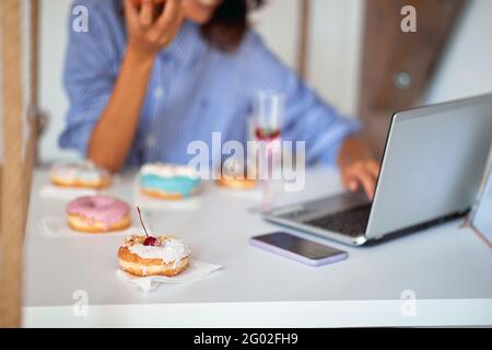 Eine Frau isst in einer angenehmen Atmosphäre in einer Konditorei leckere Donuts, während sie sitzt und einen Laptop benutzt. Konditorei, Dessert, süß Stockfoto