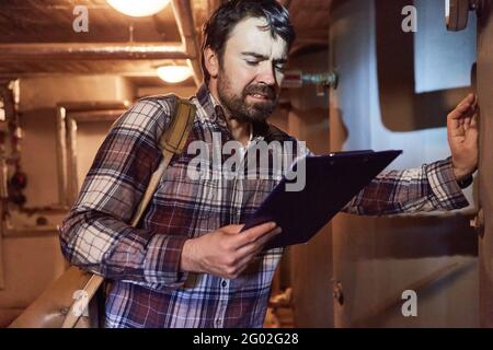 Der Heizungsinstalleur überprüft den Brennwertkessel einer Gasheizung Im Untergeschoss mit einer Checkliste Stockfoto