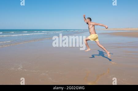 Junger Hipster-Mann mit Bart im gelben Badeanzug läuft und springt an einem einsamen Strand ins Meer. Freizeit- und Urlaubskonzept Stockfoto