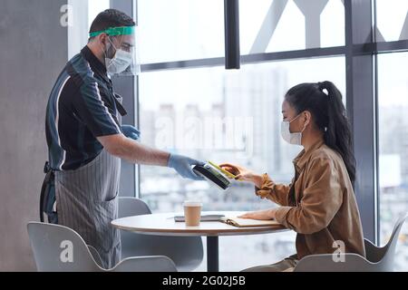 Seitenansicht Porträt eines Kellners mit Gesichtsschutz und Handschuhen Während der Betreuung weiblicher Gäste im Café mit kovidierten Sicherheitsmaßnahmen Stockfoto