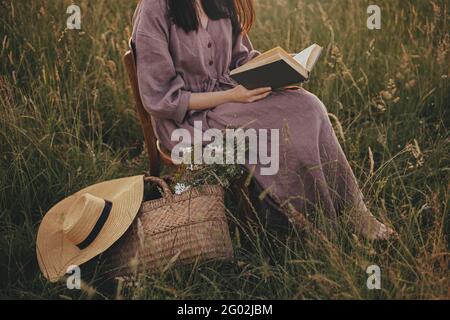 Schöne Frau in Leinenkleid mit Buch und Korb mit Blumen sitzen auf Holzstuhl in Sommerwiese, Nahaufnahme. Junge Frau, die sich abends in Co. Entspannt Stockfoto