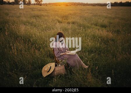 Schöne Frau in Leinenkleid mit Buch und Korb mit Blumen sitzen auf Holzstuhl im Sommer Wiese in Sonnenuntergang. Junge Frau, die sich abends in Co. Entspannt Stockfoto