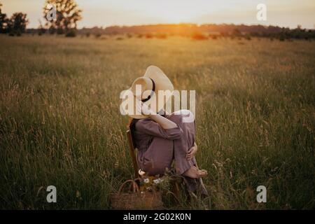 Schöne Frau in Leinenkleid und Strohhut sitzen auf rustikalem Stuhl in Sommerwiese in Sonnenuntergang. Junges Weibchen mit Blumenkorb, das sich in der Zählung entspannt Stockfoto