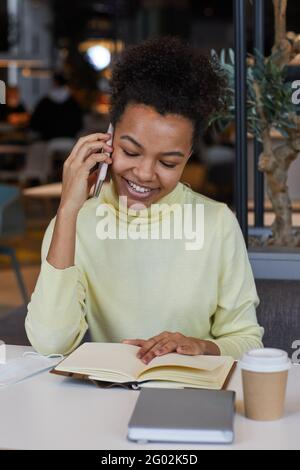 Vertikales Porträt einer lächelnden afroamerikanischen Frau, die mit dem Smartphone spricht Studieren oder Arbeiten am Kaffeetisch Stockfoto